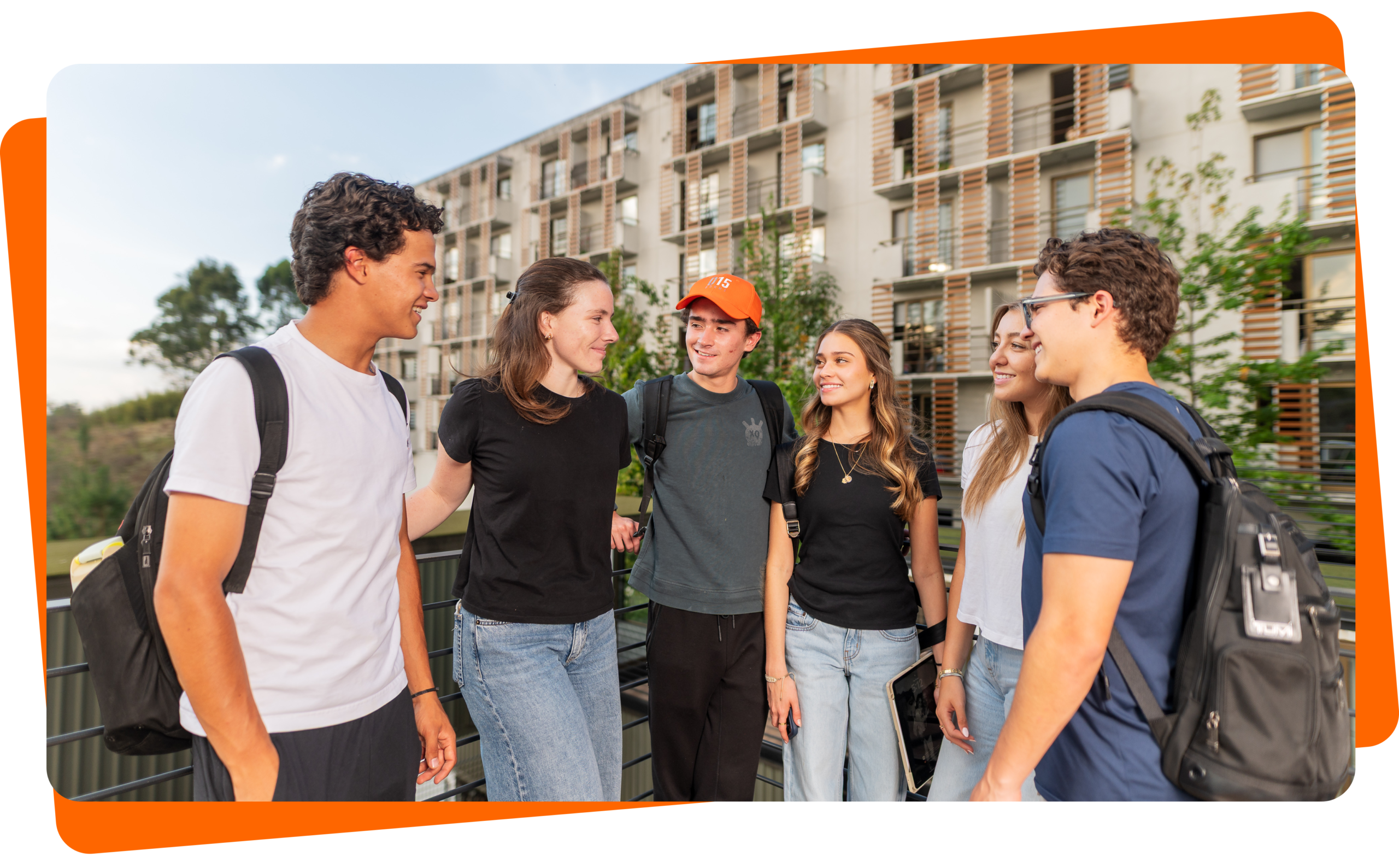 Grupo de seis jóvenes universitarios con mochilas conversando en la terraza de los edificios modernos de D15 Dorms.